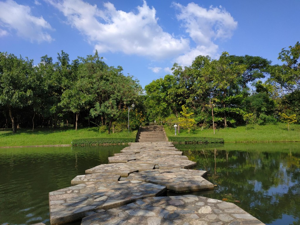 Stone stepping blocks cross calm water toward stairs and leafy trees.