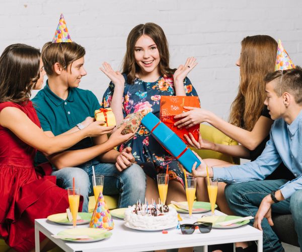 Partygoers present wrapped gifts to a smiling woman beside a table with a birthday cake and drinks.