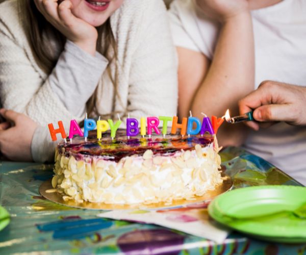 A hand lights colorful “HAPPY BIRTHDAY” candles on a cake with people seated behind it.