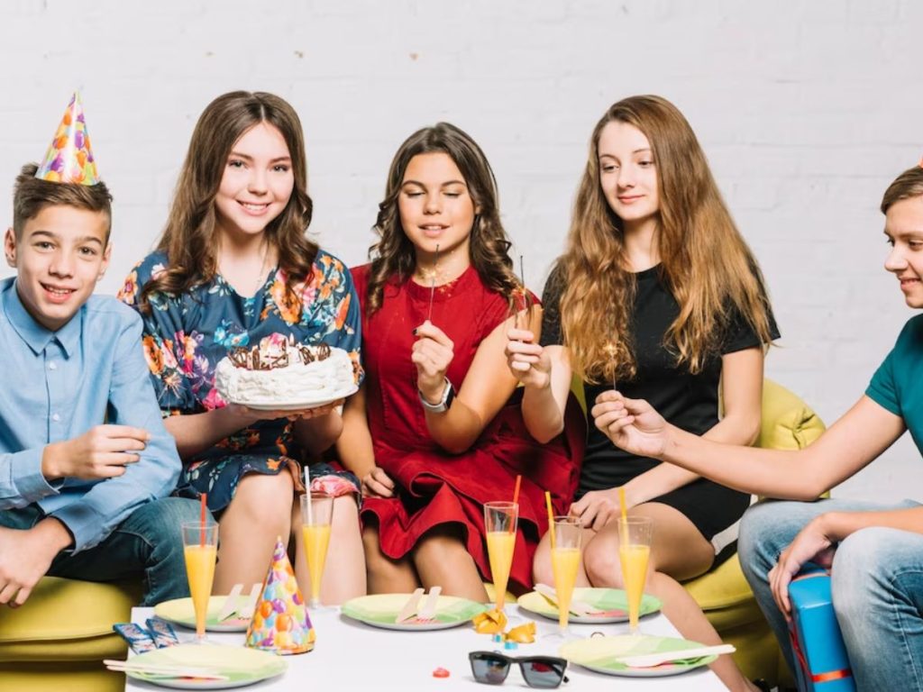 Young people celebrate with a white-frosted cake and sparklers beside a table set with drinks and party hats.