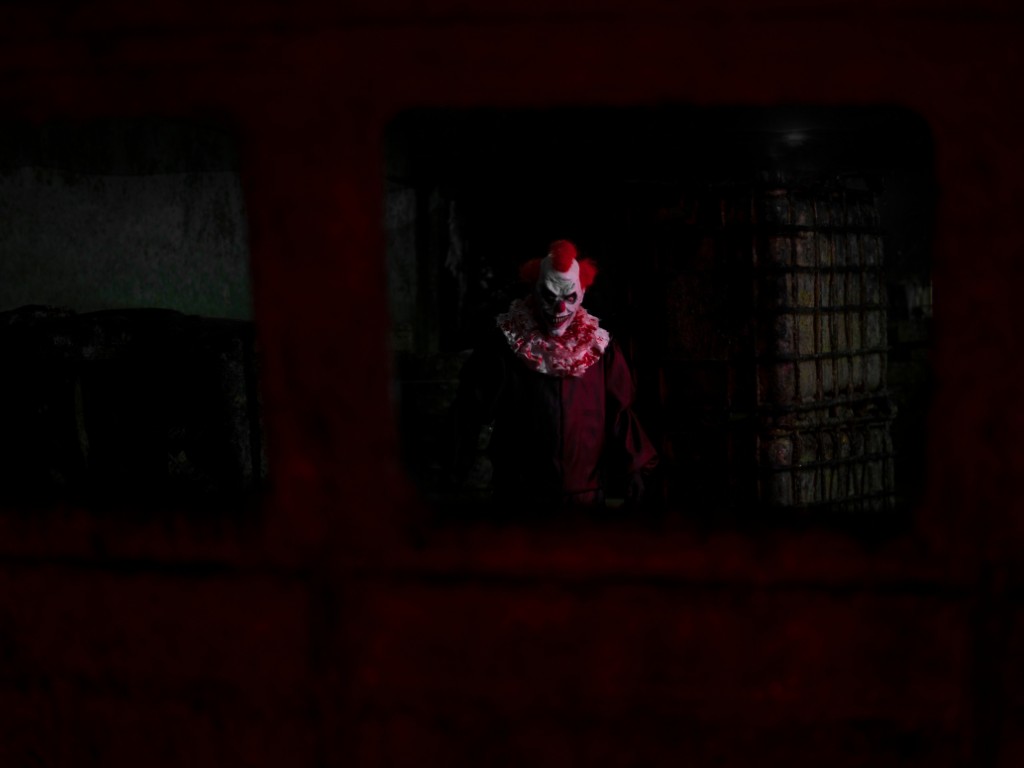 Sinister red-haired clown standing in a dark room, seen through a barred opening.