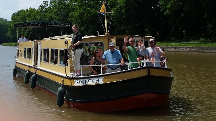 Passengers waving from the yellow-and-black Sam Patch boat on a tree-lined canal