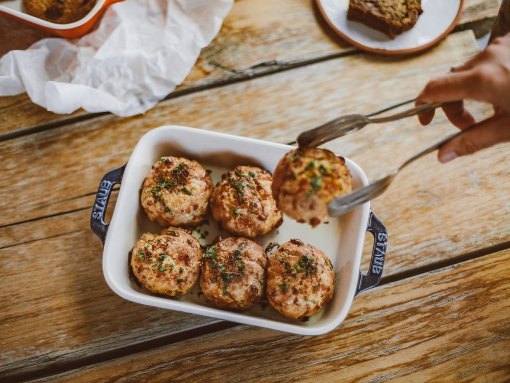 Tongs lift a golden-brown, herb-topped bite from a white baking dish.