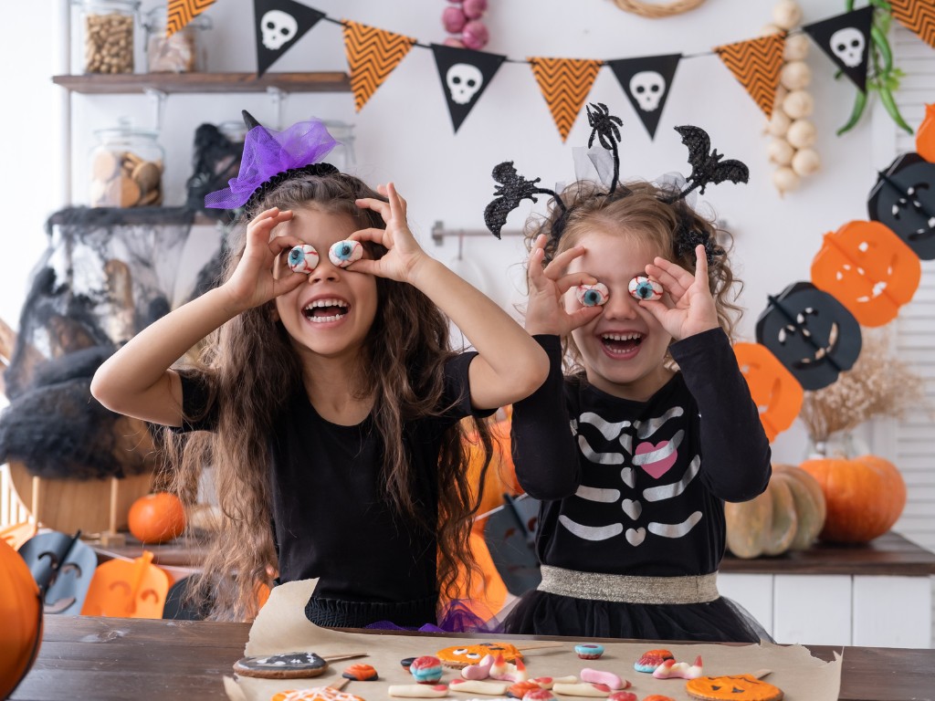 Two children in Halloween costumes hold eyeball-shaped treats over their eyes beside a table of decorated cookies.