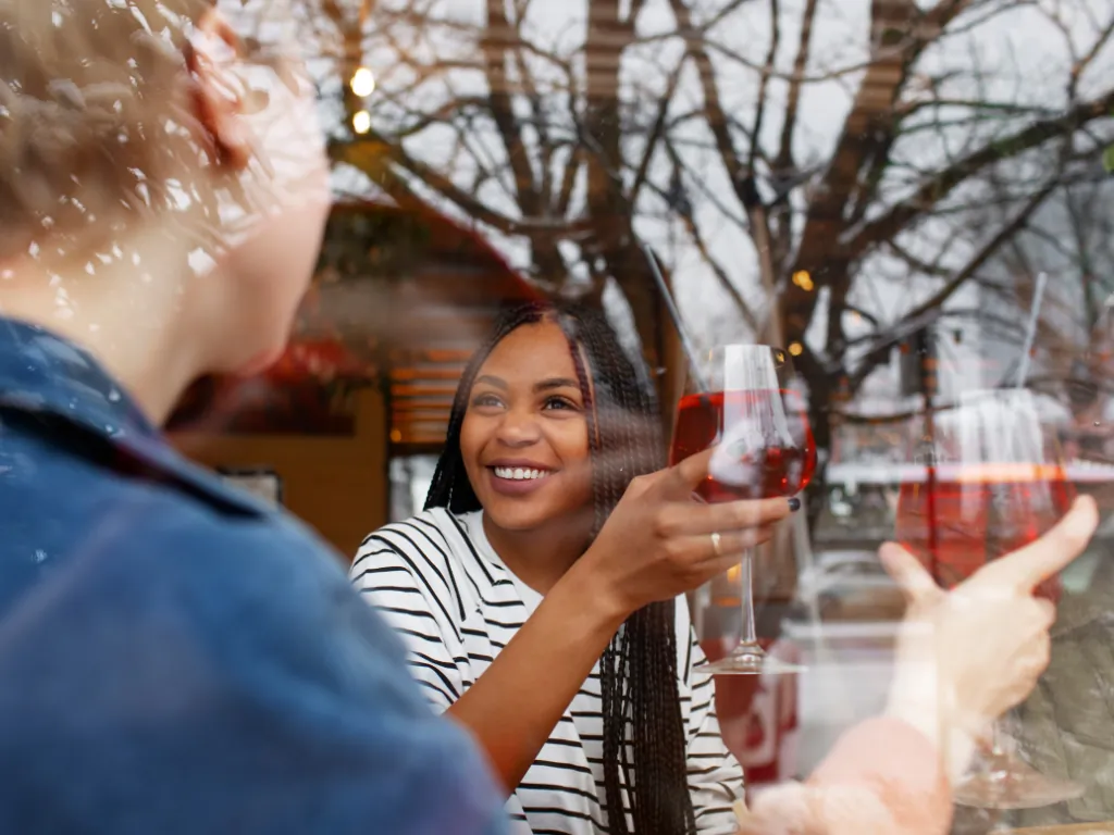 Two people raise glasses of reddish wine, seen through a window reflecting tree branches.