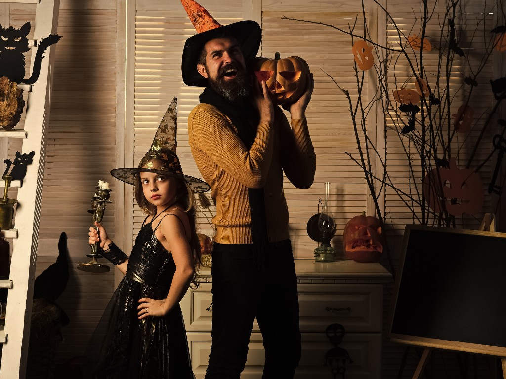 An adult and child in witch hats holding a jack-o’-lantern and candlestick among Halloween decorations.