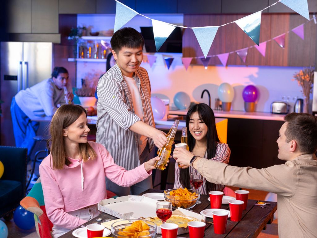 Partygoers toasting around a kitchen table with pizza, chips and drinks