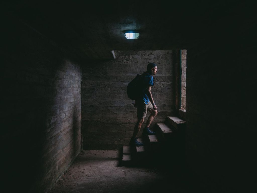 A person wearing a backpack climbs a dim concrete stairwell toward a brighter opening.