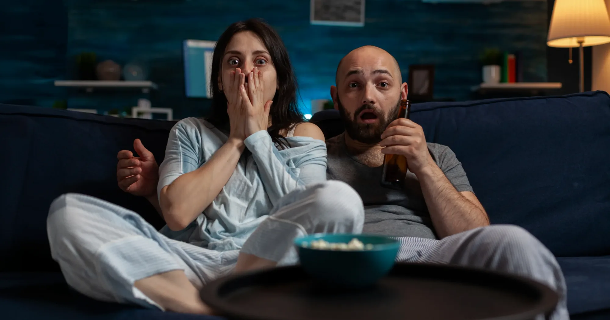 Wide-eyed couple on a couch at night with a bowl of popcorn, one covering their mouth in fright