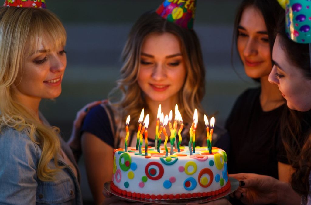Four women gathered around colorful birthday cake with lit candles
