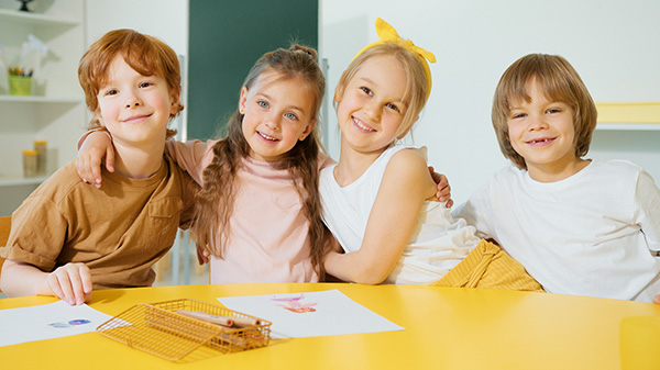 Four smiling children posing together at a table with drawings and pencils
