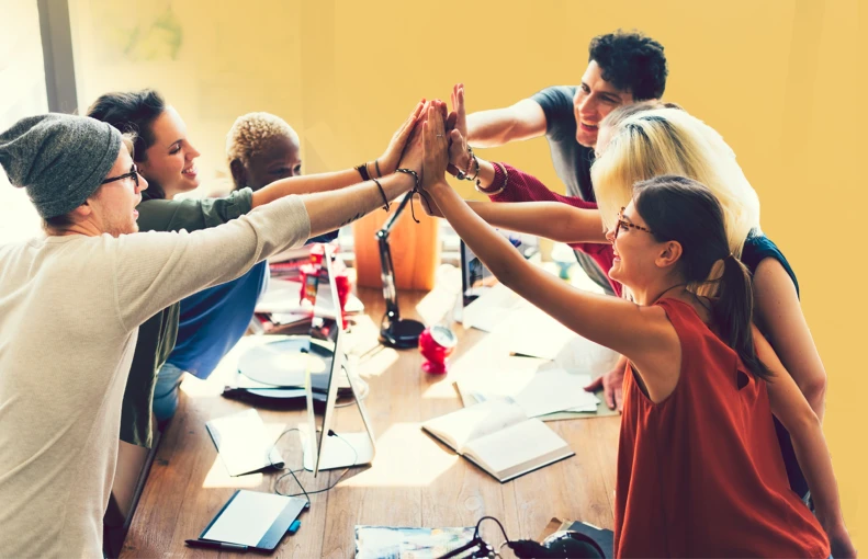 Colleagues join hands for a group high-five around a table.